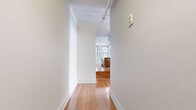 This single-floor urban residence combines contemporary finishes with classic architectural character throughout its well-designed layout. Warm-toned hardwood flooring flows seamlessly from room to room, providing a cohesive and inviting foundation across the entire space.

The kitchen is a standout feature, appointed with rich, dark wood cabinetry that extends to the ceiling, offering ample storage. A generous central island with a double sink and light countertops serves as both a functional workspace and a social hub. Stainless steel appliances, including a gas range, microwave, and dishwasher, complete the well-equipped kitchen. Large double-hung windows with green-trimmed frames frame views of an ornate neighboring facade, filling the kitchen with natural light.

The open living area is spacious and airy, with multiple large windows along the corner walls that invite an abundance of natural light and offer dynamic urban views of the surrounding cityscape. Exposed industrial-style ductwork adds a loft-like character to the space.

A corridor with a track lighting system and rail connects the living areas to the private quarters, where the bathroom features a clean, tiled soaking tub and shower combination with chrome fixtures. Throughout the space, crisp white walls and generously sized baseboards contribute to a bright, modern aesthetic with an urban edge.