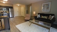 Manchester Unit 2109 is a single-floor residence designed with a practical and comfortable layout. The living area serves as the heart of the unit, showcasing smooth wood-style flooring that extends throughout the open space, lending warmth and visual continuity to the interior. Neutral-toned walls provide a versatile backdrop, allowing the space to feel bright and adaptable to various décor styles.

The living area is generously proportioned, offering ample room for seating arrangements and everyday furnishings. A glass-top coffee table and a patterned accent chair complement the space alongside a dark sofa, illustrating the room's capacity to accommodate comfortable, functional furniture groupings. Ceiling-mounted lighting provides consistent illumination throughout.

The entryway is clearly defined, featuring white paneled doors that contribute to a clean, classic aesthetic. A utility panel is conveniently recessed into the wall near the entry, maintaining a tidy appearance. An additional doorway visible from the living area hints at further rooms beyond. Overall, the unit presents a straightforward, well-maintained interior well-suited for comfortable everyday living.