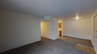 This single-floor residence offers a straightforward, functional layout designed to maximize usable living space. The main living area is generously sized, finished with wall-to-wall carpet that adds warmth underfoot and a neutral, inviting tone throughout. Crisp white walls and a smooth white ceiling keep the interior feeling bright and clean, while HVAC vents are neatly integrated into the ceiling for efficient climate control.

A central architectural wall divides the main living space, creating a natural sense of separation between distinct areas while maintaining an open feel. This structural element adds subtle definition to the floor plan without fully closing off any section of the space.

Through an opening in the dividing wall, a glimpse into the kitchen reveals warm wood cabinetry that introduces a contrast of natural tones against the otherwise neutral palette. The kitchen appears compact yet well-equipped for everyday use, with cabinetry providing ample storage.

Overall, the space presents a clean, unfussy aesthetic with practical finishes throughout. The combination of neutral tones, carpeted floors, and connected living and kitchen areas creates a cohesive and livable environment suited for comfortable day-to-day living.