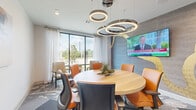 Located at 1724 Partnership Way in Houston, TX, this contemporary conference room offers a polished and professional environment designed for productive meetings and collaboration. The centerpiece of the room is a round wooden table surrounded by ergonomic orange and neutral-toned chairs, creating an inclusive and functional seating arrangement. Overhead, a striking set of cascading circular ring pendant lights serves as a bold design statement while providing ample illumination throughout the space. One wall features textured gray tile paneling adorned with an abstract gold brushstroke mural, adding an artistic focal point complemented by a large wall-mounted flat-screen display. Floor-to-ceiling windows dressed with sheer white curtains flood the room with natural light and offer views of the surrounding greenery, enhancing the bright and open atmosphere. A cozy lounge corner near the windows includes accent chairs and a small side table, providing a more relaxed seating option. The flooring combines layered patterned carpet tiles in earthy tones, contributing texture and visual interest. Glass sliding doors with black frames connect the room to the broader facility, maintaining visual openness while ensuring privacy when needed. Decorative touches such as potted plants and contemporary wall art complete the sophisticated, modern aesthetic of this well-appointed meeting space.