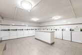 Located at 136 Bay St S in Hamilton, Ontario, this shared laundry facility offers a clean, efficiently organized space designed for practical, everyday use. The room is finished with large-format white ceramic wall tiles accented by a continuous horizontal taupe tile border, lending the space a tidy, uniform aesthetic. Light grey floor tiles run throughout, contributing to the bright and sanitary atmosphere.

A row of top-loading washing machines lines one wall, accompanied by a corresponding bank of front-loading dryers along the adjacent wall, providing ample laundry capacity for building residents. A centrally positioned white countertop island offers a convenient surface for sorting and folding laundry. Flush-mounted ceiling light fixtures ensure consistent, even illumination throughout.

Utility infrastructure, including exposed overhead piping, is neatly routed along the ceiling. Additional amenities include a coin-operated payment kiosk, wall-mounted soap and paper towel dispensers, a utility sink, waste and recycling receptacles, and built-in bench seating along the walls for resident comfort. A fire extinguisher and safety signage are also present, reflecting attention to building safety standards. The space is practical, well-maintained, and thoughtfully outfitted to serve the needs of building occupants efficiently.