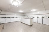 Located at 136 Bay St S in Hamilton, Ontario, this shared laundry facility offers a clean, efficiently organized space designed for practical, everyday use. The room is finished with large-format white ceramic wall tiles accented by a continuous horizontal taupe tile border, lending the space a tidy, uniform aesthetic. Light grey floor tiles run throughout, contributing to the bright and sanitary atmosphere.

A row of top-loading washing machines lines one wall, accompanied by a corresponding bank of front-loading dryers along the adjacent wall, providing ample laundry capacity for building residents. A centrally positioned white countertop island offers a convenient surface for sorting and folding laundry. Flush-mounted ceiling light fixtures ensure consistent, even illumination throughout.

Utility infrastructure, including exposed overhead piping, is neatly routed along the ceiling. Additional amenities include a coin-operated payment kiosk, wall-mounted soap and paper towel dispensers, a utility sink, waste and recycling receptacles, and built-in bench seating along the walls for resident comfort. A fire extinguisher and safety signage are also present, reflecting attention to building safety standards. The space is practical, well-maintained, and thoughtfully outfitted to serve the needs of building occupants efficiently.