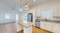 This single-story home presents a clean, modern aesthetic defined by crisp white walls, light hardwood-style flooring, and an abundance of natural light throughout. The open-concept kitchen serves as the heart of the home, showcasing white shaker-style cabinetry, gray quartz countertops, and a full suite of stainless steel appliances including a refrigerator, electric range, microwave, and dishwasher. A central island with pendant lighting overhead provides additional workspace and casual dining potential. Multiple windows with white blinds allow generous natural light to fill the kitchen and adjoining living areas, both of which are equipped with ceiling fans.

The living spaces are generously proportioned with clean lines and neutral tones, creating a versatile, move-in-ready environment. Bedrooms continue the home's consistent aesthetic, featuring the same light hardwood flooring, ceiling fans, and white walls. Closet storage is available within the bedroom areas. The bathrooms include a tiled tub-shower combination with brushed gold fixtures in one, and a separate glass-enclosed walk-in shower with a vanity and natural light in another. The overall design is cohesive, modern, and bright.
