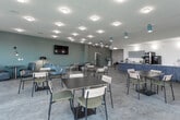 Located along I-29 North in North Kansas City, Missouri, this single-floor communal space combines café-style dining, relaxed lounge seating, and bright, open gathering areas into one cohesive environment. The interior is finished with polished concrete flooring and soft teal-painted walls, creating a calm, contemporary atmosphere throughout.

The space is divided into distinct zones. A beverage bar anchors one side of the room, featuring a long counter with textured blue wood-grain cabinetry, open display shelving with white tile backsplash, and professional coffee and beverage equipment. Adjacent to the bar, clustered café tables with mixed wood-and-metal chairs provide flexible dining and casual work seating. Recessed ceiling lights and teal pendant fixtures illuminate the space evenly.

A dedicated lounge area offers a full sofa, a modern accent chair, a layered round coffee table, and a wall-mounted flat-screen television, complemented by a statement floor lamp. Intimate booth seating with upholstered banquettes and articulating wall sconces creates a quieter nook nearby.

On the opposite end, floor-to-ceiling windows and glass doors flood the dining area with natural light and provide unobstructed views of the surrounding outdoor greenspace, enhancing the open, airy quality of the overall space.