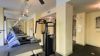 This fitness center offers a practical and well-appointed space designed to support a full workout experience. The single-floor facility is outfitted with a selection of strength training equipment, including a multi-function cable machine, as well as cardio machines such as rowing machines and treadmills visible in the background. The layout is thoughtfully organized, with structural columns defining workout zones without obstructing the open flow of the space. Walls are finished in warm neutral tones, contributing to an inviting and energizing atmosphere. Dark rubberized flooring provides both safety and durability underfoot, ideal for high-traffic fitness use. Track lighting and ceiling fans are installed throughout, ensuring the space remains well-lit and properly ventilated during use. Large mirrored panels line portions of the walls, enhancing the sense of openness while allowing users to monitor their form. A water dispenser is also visible, adding a convenient amenity for those working out. The overall design balances functionality with comfort, making this facility a well-rounded option for daily fitness routines.
