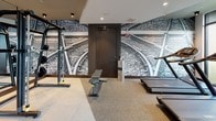 Located at 88 Cambridgepark Dr in Cambridge, MA, this professionally appointed fitness center delivers a comprehensive workout environment with a sleek, contemporary design. The space is organized into distinct functional zones, seamlessly blending cardio and strength training areas within a single, open floor plan.

The cardio zone is lined with floor-to-ceiling windows that flood the space with natural light and offer views of the surrounding greenery, creating an energizing atmosphere for treadmill and elliptical workouts. A row of Technogym cardio machines — including treadmills, ellipticals, and stair climbers — are positioned to take full advantage of the outdoor views.

The strength training area features a broad selection of Technogym selectorized machines, cable systems, free weights, and adjustable benches, all arranged to maximize space and workflow. Full-length mirrors line the walls, enhancing the sense of openness while allowing users to monitor their form. Overhead, wall-mounted televisions provide entertainment throughout workouts.

A standout design element is the dramatic black-and-white photographic mural of converging railway tracks, which spans an entire wall and adds a bold artistic focal point to the space. Recessed lighting and a neutral color palette of grays and whites lend the facility a polished, upscale character throughout.