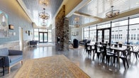 This single-story clubhouse offers an impressive open-concept layout that seamlessly integrates social and relaxation zones into one cohesive space. The most commanding feature is a floor-to-ceiling stacked stone fireplace column that anchors the room, serving as a dramatic focal point visible from nearly every angle. Soaring ceilings are adorned with decorative stamped metal tile panels, adding architectural texture and a refined industrial character throughout.

Industrial-style chandelier fixtures hang at varying heights, providing warm ambient lighting that complements the polished concrete floors below. A large ornate area rug adds warmth and definition to the central lounge area, which is furnished with plush seating arranged for comfortable gathering. Adjacent to the lounge, a dedicated dining area features sleek black metal chairs and long tables suited for communal use or events.

Expansive floor-to-ceiling windows and glass-paneled entry doors line the perimeter, flooding the interior with natural light and offering views of the surrounding exterior. The overall ambiance balances modern industrial design with inviting, comfortable furnishings, creating a welcoming environment well-suited for both casual socializing and organized gatherings.