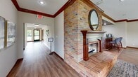 Located at 36 Broadway Village Dr in Columbia, Missouri, this single-floor space offers a thoughtfully designed layout that seamlessly combines a living room and dining area into one cohesive, welcoming environment. The space is anchored by a striking full-brick fireplace, complete with a richly detailed wood mantel, decorative columns, and a round mirror above — a classic architectural centerpiece that lends warmth and character to the room. The fireplace surround extends to brick flooring and brick wall cladding in the entryway area, creating a rustic yet refined transition into the main space. Rich wood crown molding runs along the upper perimeter of the rooms, adding a layer of traditional elegance throughout. The living area features warm hardwood-style flooring and is furnished with comfortable seating arranged around a central area rug, offering an inviting setting for gathering. The adjacent dining area is bathed in natural light, courtesy of large floor-to-ceiling windows lining multiple walls. These windows frame sweeping views of the lush, green landscape outside, bringing the outdoors in and creating a bright, airy atmosphere. The overall ambiance is both functional and comfortable, blending classic architectural details with an open, light-filled layout well-suited for communal use.