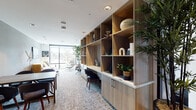 Located at 4949 Baltimore Dr in La Mesa, California, this thoughtfully designed single-floor amenity space functions as both a social lounge and a productive co-working environment. The interior showcases a contemporary aesthetic with a warm, curated palette that balances comfort and functionality throughout.

A standout feature is the expansive built-in wood shelving unit, which spans an entire wall and serves as both a practical workstation and a display area accented with decorative objects and lush greenery. Adjacent to this, a communal table with seating provides an ideal setting for collaborative work or casual gatherings.

The lounge area is anchored by a bold mustard-yellow velvet sofa paired with modern accent chairs, ottomans, and sleek gold floor lamps, creating an inviting atmosphere for relaxation. Large floor-to-ceiling windows flood the space with natural light and offer open views of the surrounding streetscape and palm-lined boulevard.

A semi-private workstation tucked beside frosted glass sliding panels offers a quieter spot for focused work, furnished with a minimalist desk and leather chair. Additional seating nooks feature a cognac leather armchair with ottoman and sculptural dark velvet chairs arranged around a solid wood table.

Textured carpeting, soft gray walls, recessed and pendant lighting, and carefully placed indoor plants complete the polished, welcoming ambiance of this versatile community space.