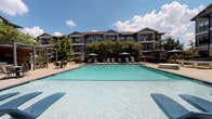 This outdoor pool area is a well-appointed amenity space designed to deliver a resort-like experience. At its center sits a generously sized swimming pool with clear, turquoise water, accompanied by a separate spa positioned nearby for relaxation. The pool deck is finished in smooth concrete and offers ample space for lounging, with rows of chaise lounge chairs arranged along the poolside and shaded by market umbrellas.

A standout architectural feature is the covered pavilion, which runs alongside the pool area and serves as a shaded gathering and lounge space. The pavilion is supported by substantial natural stone columns and finished with warm wood-panel ceiling details, ceiling fans for airflow, and large-format stone tile flooring. Glass panel railings offer unobstructed sightlines to the pool while maintaining an open, airy feel. Comfortable seating is arranged beneath the covered structure, making it an ideal spot for outdoor entertaining.

The surrounding landscaping incorporates mature trees and ornamental plantings that provide natural shade and greenery. The multi-story craftsman-influenced building forms an attractive architectural backdrop, featuring board-and-batten siding, balconies, and pitched rooflines. The overall ambiance is vibrant, social, and well-suited for outdoor leisure under open skies.