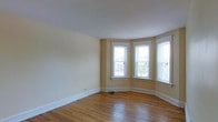 This single-floor space showcases a welcoming main room defined by its standout curved bay window alcove, which houses multiple large, white-trimmed windows offering views of the surrounding neighborhood and filling the interior with abundant natural light. The windows are thoughtfully arranged in a gentle arc, creating an architectural focal point that adds both character and brightness to the room. The flooring throughout is warm, honey-toned hardwood, lending a classic and timeless quality to the space. Walls are finished in a soft, neutral warm beige, providing a versatile backdrop suited to a variety of décor styles. Broad white baseboards run along the perimeter, adding a crisp, clean contrast to the walls and floors. The ceiling is smooth and white, contributing to an airy, open feel. The overall layout is open and unobstructed, allowing for flexible furniture arrangements. The combination of the bay window's natural light, the rich hardwood floors, and the neutral palette creates a bright, inviting ambiance with a nod to classic architectural detailing.