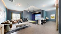 This thoughtfully designed single-floor amenity space combines leisure and functionality across a generously sized layout. The entry hallway sets an elegant tone with textured blue accent walls, warm cove lighting, large-format tile flooring, and tasteful décor including a cushioned bench and decorative console — creating a polished first impression.

The expansive living room serves as the heart of the space, featuring a sophisticated lounge area adorned with richly textured blue and grey wallcovering, a plush sectional sofa, rounded accent chairs, and a dark wood coffee table set atop a layered area rug. Natural light filters in through large windows dressed with floor-length drapes. Adjacent to the lounge, a dedicated Full Swing Golf simulator bay offers a professional-grade turf hitting mat, ceiling-mounted projector, and a large immersive screen — complemented by a high-top bar counter with seating for spectators. Golf club storage and equipment racks are neatly integrated nearby.

The office area stands out with floor-to-ceiling built-in shelving finished in deep slate blue, displaying a curated collection of kitchen appliances and accessories atop a checkered tile floor with a striking yellow ceiling detail — suggesting a versatile, well-organized utility or demonstration space. Together, these areas create a dynamic, amenity-rich environment ideal for both relaxation and recreation.