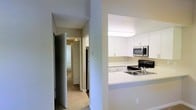 This single-floor residence presents a thoughtfully designed layout visible from a central vantage point that connects the kitchen and hallway areas. The kitchen is outfitted with bright white cabinetry extending to the upper walls, providing generous storage space. A suite of stainless steel appliances — including a gas range, built-in microwave, and refrigerator — adds a modern, functional character to the space. The countertops are finished in a clean white surface, complementing the light and airy aesthetic of the kitchen. A peninsula-style counter extends outward, offering additional workspace or casual seating potential. The sink is integrated into the counter, positioned conveniently near the range. Adjacent to the kitchen, a corridor connects to further rooms, lined with neutral-toned walls and light carpeting that lend a calm, understated atmosphere. Panel-style doors along the hallway lead to additional spaces, suggesting a well-organized floor plan. Recessed and overhead lighting throughout ensures the space remains well-lit. The overall design emphasizes practicality and cleanliness, with a neutral color palette that creates a cohesive and inviting environment throughout.