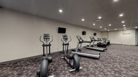 Located at 1201 E Market St in Akron, Ohio, this fitness center offers a well-rounded exercise environment designed to accommodate a wide range of workout routines. The single-floor facility features a spacious, open layout with patterned carpet flooring throughout and a drop tile ceiling with recessed lighting that keeps the space well-illuminated.

The cardio section is generously equipped with multiple treadmills, elliptical trainers, upright and recumbent stationary bikes, and a stair climber — each station conveniently positioned beneath wall-mounted flat-screen televisions. A dedicated strength training area includes a multi-function cable machine, a full dumbbell rack with a variety of weight options, a weight bench, and a medicine ball rack. A large wall mirror in the strength zone enhances spatial awareness and supports proper form during workouts.

An open stretch and functional training area provides room for floor exercises, with stability balls and exercise mats available for use. Accent walls in teal blue add a pop of color against the otherwise neutral gray and white palette, contributing to an energizing yet focused atmosphere. The facility is accessible via double doors and includes a water cooler station, ensuring convenience throughout your workout.