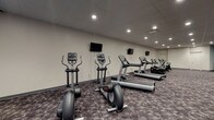 Located at 1201 E Market St in Akron, Ohio, this fitness center offers a well-rounded exercise environment designed to accommodate a wide range of workout routines. The single-floor facility features a spacious, open layout with patterned carpet flooring throughout and a drop tile ceiling with recessed lighting that keeps the space well-illuminated.

The cardio section is generously equipped with multiple treadmills, elliptical trainers, upright and recumbent stationary bikes, and a stair climber — each station conveniently positioned beneath wall-mounted flat-screen televisions. A dedicated strength training area includes a multi-function cable machine, a full dumbbell rack with a variety of weight options, a weight bench, and a medicine ball rack. A large wall mirror in the strength zone enhances spatial awareness and supports proper form during workouts.

An open stretch and functional training area provides room for floor exercises, with stability balls and exercise mats available for use. Accent walls in teal blue add a pop of color against the otherwise neutral gray and white palette, contributing to an energizing yet focused atmosphere. The facility is accessible via double doors and includes a water cooler station, ensuring convenience throughout your workout.