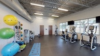 This fitness center offers a spacious, single-floor layout designed to accommodate a wide range of workout routines. The facility features soaring ceilings with an exposed steel truss structure and industrial ductwork, lending the space an open, warehouse-style aesthetic. Rows of fluorescent lighting ensure the entire floor is well-lit throughout the day.

The cardio zone lines the windowed wall, where multiple elliptical trainers, a recumbent bike, and treadmills are arranged to take advantage of the natural light and outdoor views provided by the large windows. Wall-mounted flat-screen televisions are positioned throughout this area for entertainment during workouts.

The strength and functional training area is equally well-appointed. A multi-station cable machine, a dumbbell rack with an adjustable bench, and a dedicated weight bench provide a solid foundation for resistance training. A colorful stability ball rack, medicine balls, resistance bands, and yoga mats support flexibility and functional fitness routines, with ample open floor space available for stretching or bodyweight exercises.

The durable rubber flooring covers the entire workout area, offering both safety and comfort. A glazed interior door leads to an adjacent room, and the clean white walls contribute to a bright, motivating atmosphere throughout the center.
