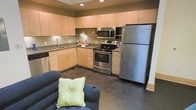 This single-floor space features an open-concept layout that seamlessly connects the kitchen and living area, creating a functional and cohesive environment. The kitchen is well-appointed with a full suite of stainless steel appliances, including a refrigerator, electric range, built-in microwave, and dishwasher. Light maple cabinetry lines the walls in an L-shaped configuration, offering generous storage from floor to ceiling. Dark granite-style countertops provide a contrasting work surface, while a stainless steel sink with a window backsplash adds both practicality and brightness. Recessed lighting illuminates the kitchen area effectively. The dark tile flooring extends throughout, grounding the space with a bold, contemporary feel. The adjoining living area accommodates comfortable seating, as seen with the navy blue sofa accented by a light green accent pillow, reinforcing the casual and functional character of the space. The open layout between the kitchen and living zone makes the overall space feel efficient and well-utilized for everyday living.