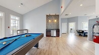 Located at 345 Buckland Hills Drive in Manchester, Connecticut, this single-story community clubhouse offers a well-appointed gathering space designed for recreation and socializing. The open-concept layout is anchored by soaring vaulted ceilings and bathed in natural light from multiple sets of French doors and large transom windows that frame views of the surrounding greenery.

The main game area is a standout feature, equipped with a full-size pool table and a foosball table, both set atop light-toned hardwood flooring that runs throughout the space. A built-in wet bar with dark cabinetry, a stainless steel appliance, and a striking mosaic tile backsplash serves as a focal point, illuminated by pendant lighting overhead.

Adjacent to the game room, a fireplace lounge provides a relaxed gathering spot, flanked by two sets of French doors that open to the outdoors, creating a bright and airy atmosphere. A ceiling fan adds to the comfort of the space. A wall-mounted flat-screen television and decorative columns add architectural interest throughout.

A cozy nook with a high-top bistro table and bar-height seating beside a large window offers an intimate spot for casual conversation. White trim and soft blue-gray walls lend the entire clubhouse a clean, modern, and inviting aesthetic.