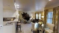 This single-floor residence offers an inviting open-concept layout that seamlessly connects the kitchen, dining, and living areas into one cohesive and functional space. The living room serves as the heart of the home, anchored by a striking cowhide wall accent above the media console, which adds a bold, eclectic touch to the otherwise warm and neutral palette. A plush, carpeted floor runs throughout the main living area, contributing to the cozy ambiance. Comfortable armchairs in a soft teal upholstery are arranged around a petite round side table, while a rich brown leather sofa and accent chair round out the seating arrangement near the window. Elegant gold and cream patterned curtains frame the natural light filtering through the windows, enhancing the warm tones of the room. The dining area features a round glass-top table with ornate metal legs, surrounded by patterned upholstered chairs — a refined centerpiece accented by a fresh floral arrangement. Adjacent to the living space, the kitchen is equipped with white cabinetry and a raised breakfast bar with stylish bar stools, offering casual seating and a natural visual separation between the kitchen and the main living area. Recessed lighting throughout ensures the space is well-lit and welcoming.