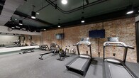 Located at 1218 Izard St in Omaha's historic district, this fitness center blends industrial character with modern functionality. The single-floor exercise room is defined by striking exposed brick walls, original structural wood columns, and a dark ceiling with visible ductwork and piping — details that lend the space a distinctive loft-like aesthetic.

The flooring throughout is durable rubber gym tile, providing a safe and practical surface for all types of workouts. Cardio equipment includes multiple treadmills and air bikes, while strength training is supported by a cable machine, dumbbell rack with bench, and a variety of stability and medicine balls mounted on the wall. Large wall-mounted flat-screen televisions are positioned throughout the space for entertainment during workouts.

A full-length mirror along one wall enhances the sense of openness, and recessed and pendant lighting ensures the space is well-lit. A water station and additional amenity fixtures are accessible near the entrance, which features glass partition doors. The layout is thoughtfully organized to accommodate both cardio and functional training, making this a comprehensive and well-appointed fitness facility.