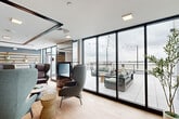 Located in Chelsea, MA, the Sky Lounge is a thoughtfully designed rooftop amenity space that seamlessly blends interior comfort with expansive outdoor living. The interior lounge area features light hardwood flooring, floor-to-ceiling black-framed glass walls, and a striking double-sided fireplace that serves as a dramatic focal point. Carefully selected seating, including high-back accent chairs and intimate conversation groupings, creates a welcoming and sophisticated atmosphere throughout. An abundance of natural light pours in through the expansive glass façade, offering unobstructed sightlines to the outdoor terrace and beyond. The wet bar area is outfitted with warm wood cabinetry, dark countertops, a large center island with bar seating, and a sculptural three-dimensional tile accent wall, all illuminated by pendant lighting for a modern yet inviting feel. The outdoor terrace is generously proportioned, finished with large-format porcelain tile and furnished with plush seating arrangements accented by string lights overhead. From the terrace, sweeping views of the Boston skyline and the surrounding waterfront create a striking backdrop, making this rooftop lounge an exceptional space for gathering, relaxing, or entertaining.