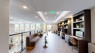 This well-appointed amenity lounge is designed to serve as a sophisticated communal gathering and work space. The interior features light hardwood-style flooring throughout, paired with a striking tray ceiling outfitted with recessed lighting that creates a warm, layered ambiance. Along one wall, dark wood built-in shelving units and a dedicated workstation area with bar-height seating provide a functional co-working environment, complete with a desktop computer and decorative displays. On the opposite side, a sleek curved sofa with a gold base accent anchors a casual lounge area, accompanied by pedestal side tables in a brushed bronze finish. Large windows along the exterior wall flood the space with abundant natural light and offer views of the surrounding outdoor terrace and palm-lined courtyard beyond. A modern cable-rail balustrade lines one edge of the interior, hinting at the multi-level nature of the broader building. The adjacent outdoor terrace extends the living space further, featuring comfortable lounge chairs, modular seating arrangements, large planters with tropical palms, and direct views of a resort-style pool. The overall aesthetic blends contemporary design with warm, residential touches, creating a welcoming environment suited for both relaxation and productivity.