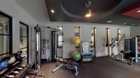 This single-floor fitness center is thoughtfully designed to support a comprehensive workout experience across multiple distinct training zones. The space is immediately distinguished by its striking tray ceilings, which are finished in bold accent colors — deep charcoal and rich red — and fitted with recessed lighting and pendant fixtures that create a dynamic, energizing ambiance throughout.

The cardio zone features a row of treadmills and elliptical trainers positioned along a wall of large, floor-to-ceiling windows, allowing natural light to pour in and offering views of the surrounding outdoor environment. The open layout ensures generous floor space between machines, promoting easy movement and accessibility.

The strength training area is equally well-appointed, housing a multi-cable functional trainer, a full rack of dumbbells in graduated weights, colorful kettlebells, and a variety of resistance and selectorized weight machines. Stability balls and accessories are neatly organized alongside benches, providing options for both guided and free-form training.

Wall-mounted mirrors are strategically placed throughout, amplifying the sense of space and allowing users to monitor their form. The durable rubber flooring underfoot provides both safety and noise reduction. The combination of purposeful layout, bold architectural detailing, and abundant natural light makes this fitness center a functional and visually engaging space.
