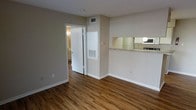 This single-floor space offers a clean, functional layout with a cohesive neutral color palette throughout. The main living area is finished with warm hardwood-style flooring that adds character and continuity across the space. Smooth, taupe-toned walls create a calm and welcoming ambiance, complemented by white trim and baseboards for a crisp contrast.

A notable architectural feature is the pass-through window connecting the kitchen to the living area, topped with a stone-style counter and framed by a wall-mounted range hood — a practical detail that enhances the sense of openness between the two spaces. The kitchen is visible through the opening, hinting at a compact but purposeful cooking area.

Ceiling-mounted HVAC vents and a wall-mounted utility panel indicate that climate control is integrated throughout the unit. An open doorway leads to what appears to be a bathroom, where warm lighting and matching flooring are visible, maintaining a consistent design language throughout the space.

Overall, this space presents a straightforward, well-kept interior that balances practicality with a tidy, move-in-ready aesthetic. The uniform flooring, neutral tones, and functional pass-through design make it a versatile and comfortable environment.