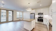 Located in Austin, TX, this well-designed one-bedroom, one-bathroom residence offers a clean, modern aesthetic with an efficient and functional layout. The open-concept kitchen and living area forms the heart of the space, anchored by a central island with a warm-toned quartz countertop and crisp white cabinetry. The kitchen is fully equipped with a full suite of stainless steel appliances, including a refrigerator, range, microwave, and dishwasher, complemented by a white subway tile backsplash and recessed lighting overhead. A statement pendant light fixture adds a refined touch above the island. Light wood-style flooring runs consistently throughout the unit, lending a bright, cohesive feel. The living area features French doors and large windows fitted with blinds, filling the space with natural light. The bedroom is a comfortable size, complete with a ceiling fan, ample natural light from a large window, and access to a dedicated closet. The bathroom is outfitted with large-format white tile flooring, a tub-shower combination with white tile surround, a vanity with matching cabinetry, and built-in open shelving for additional storage. Neutral wall tones and recessed lighting throughout contribute to the space's bright, contemporary ambiance.