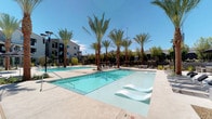This expansive outdoor amenity space is designed with a resort-style aesthetic, offering a comprehensive range of leisure and entertainment features. The centerpiece is a generously sized swimming pool with crystal-clear turquoise water, flanked by in-water lounge chairs and surrounded by a wide concrete deck lined with tall palm trees that lend a tropical ambiance. Adjacent to the main pool is a dedicated spa, tiled in a complementary palette and equipped with built-in seating, with additional lounge chairs positioned nearby for relaxation.

A fire pit lounge area provides an inviting gathering spot, furnished with woven rattan chairs and plush white cushions arranged around a sleek rectangular gas fire table. Nearby, a partially enclosed pergola seating area features matching outdoor sofas and a mirrored coffee table, framed by climbing greenery for a shaded retreat. The outdoor kitchen station is well-appointed with a stainless steel grill, utility sink, and stone-clad countertop, ideal for outdoor entertaining. A covered dining pavilion completes the space, offering multiple tables and chairs beneath a dramatic oversized ceiling fan, with a wall-mounted television making it functional for gatherings. Desert-adapted landscaping ties the entire space together.