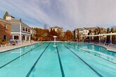 This outdoor amenity space is centered around a generously sized swimming pool with clearly marked lap lanes, making it suitable for both recreational and fitness swimming. The pool's vivid turquoise water is complemented by a wide concrete deck lined with blue lounge chairs and a white pergola structure, providing shaded seating options. Lush mature trees form a natural backdrop along the perimeter fence, enhancing the sense of privacy and creating a serene outdoor atmosphere.

Adjacent to the pool is a covered pavilion with a single-level layout that serves multiple functions. One section features a ping-pong table positioned beneath a ceiling fan, with colorful Adirondack chairs adding a casual, welcoming touch. Large arched openings along one wall frame unobstructed views of the pool and surrounding greenery, flooding the space with natural light and creating a seamless indoor-outdoor connection. Tile flooring runs throughout the covered areas, maintaining a clean and cohesive aesthetic.

An additional covered area provides outdoor dining with a table and chairs, ideal for social gatherings. Restroom facilities are accessible directly from the pavilion, and a prominently posted pool rules sign ensures clear communication for all users. The overall design balances functionality with comfort, offering a comprehensive and inviting aquatic recreational destination.