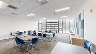 Located at 431 Church St in Norfolk, VA, this contemporary classroom or collaborative meeting space offers a thoughtfully designed, single-room layout that comfortably accommodates group learning and collaboration. The room is furnished with multiple modular rectangular tables paired with blue stackable chairs, allowing for flexible seating arrangements to suit a variety of instructional or meeting formats. Patterned, multi-tone carpeting covers the floor throughout, adding both visual interest and acoustic comfort underfoot. A standout design feature is the bold, blue-and-white checkered acoustic panel installation mounted on one wall, which serves both a functional and aesthetic purpose. Recessed fluorescent lighting in a drop-tile ceiling provides bright, even illumination across the entire space. Large floor-to-ceiling windows along one wall flood the room with natural light and offer open views of the surrounding Norfolk streetscape. A wall-mounted flat-screen display and a credenza unit are positioned along one wall, supporting presentations and storage needs. The overall ambiance is clean, professional, and contemporary, making this space well-suited for educational instruction, team meetings, or training sessions.