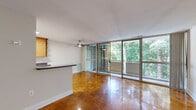 This 1-bedroom, 1-bathroom unit offers a thoughtfully organized single-floor layout. The combined living and kitchen area forms the heart of the space, immediately drawing attention to its standout feature: a full wall of floor-to-ceiling sliding glass doors that open onto a private balcony overlooking a canopy of mature trees. The abundant natural light filtering through the greenery creates a serene, nature-connected ambiance. Polished parquet flooring in a warm honey tone runs throughout the living, dining, and bedroom areas, adding a classic architectural detail. A ceiling fan anchors the living room, and a white brick accent wall adds textural contrast.

The kitchen is separated by a breakfast bar and features warm wood cabinetry with open shelving, dark granite-style countertops, white tile flooring, and a full suite of black appliances including a gas range, refrigerator, and dishwasher. The bedroom is a comfortable, well-proportioned retreat with a large window framing tree views and access to a closet. The bathroom includes a full tub-shower combination with clean white tile surround, a toilet, and a vanity.