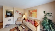 This single-floor residence offers a thoughtfully organized layout with distinct living spaces that flow naturally together. The kitchen is efficiently designed as a galley-style space, featuring dark wood cabinetry, light speckled countertops, a stainless steel sink, and black appliances including an electric range. Wood-look flooring adds warmth to the kitchen's practical design.

The living room is a welcoming space finished with carpet flooring and neutral beige walls. A large window dressed with sheer curtains and valances allows natural light to brighten the room, while ample wall space accommodates artwork. The dining area, finished with hardwood-style flooring, comfortably fits a four-seat high-top table and is accented by a wall mirror and coordinating artwork.

The bedrooms feature plush carpet, ceiling fans with lighting fixtures, and windows dressed with layered curtains that provide both privacy and natural light. One bedroom includes a built-in vanity area, adding a convenient functional touch. Throughout the home, a cohesive warm color palette of beige, terracotta, and earthy tones creates a consistently inviting and comfortable ambiance.