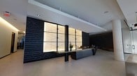 This contemporary lobby space makes an immediate architectural statement through its carefully curated design elements. At the heart of the space sits a bold, angular reception desk finished in dark tones with a contrasting light-colored countertop, accented by a simple floral arrangement that adds a natural touch to the refined setting. Flanking the desk are dramatic backlit wall panels framed in dark metal, casting a warm, diffused glow that serves as the focal point of the room. The surrounding walls are clad in dark, textured stacked stone, providing depth and contrast against the clean white ceiling above. Recessed lighting is thoughtfully integrated throughout, supplementing the ambient illumination from the feature panels and maintaining a consistent, polished atmosphere. The flooring is finished in a smooth, light-toned material that reflects the overhead lighting, enhancing the sense of openness and scale. The overall design palette — anchored in dark charcoals, blacks, and warm neutrals — conveys a professional yet inviting environment. The space strikes a balance between minimalism and bold design, making it well-suited for a corporate office, hotel, or high-end commercial establishment.