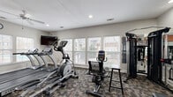 Located at 910 Saint Andrews Drive in Murfreesboro, Tennessee, this fitness center is a fully equipped exercise facility designed to support a comprehensive workout routine. The single-room space is thoughtfully arranged to maximize functionality, featuring a diverse selection of cardio equipment including True-brand treadmills, elliptical trainers, a stair climber, and stationary bikes. Strength training is well-represented with a multi-station cable machine by Inspire Fitness, a weight bench, and a dedicated free weight area with a full dumbbell rack, all reflected in a large wall-to-wall mirror that enhances the sense of space. A bold accent wall in terracotta orange adds visual character against the neutral gray walls and textured, multi-tone carpet flooring. Abundant natural light streams in through multiple white-shuttered windows along the perimeter, offering views of the surrounding community. Recessed lighting and a ceiling fan ensure the environment remains comfortable during workouts. A wall-mounted flat-screen television, a wall clock, resistance bands, stability balls, and a water station round out the amenities, making this fitness center a practical and well-appointed space for residents.