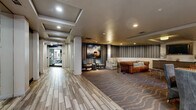 This thoughtfully designed single-floor amenity space blends contemporary style with comfortable functionality. A wide, well-lit corridor with wood-look tile flooring leads residents through the space, flanked by structural columns and recessed ceiling details that add architectural interest.

The centerpiece of the space is a generously sized lounge area anchored by a rich cognac leather Chesterfield sofa and complementary seating arrangements. Striped wallcovering in muted blue and gray tones lines the walls, creating a sophisticated backdrop alongside a bold abstract painting illuminated by a dramatic arc floor lamp. Wave-patterned carpeting grounds the seating area, adding texture and warmth.

Natural light filters in through windows dressed with patterned drapery, while decorative wall art and organic sculptural accents contribute to the curated, upscale aesthetic throughout. A dedicated game table with a felt surface and surrounding upholstered chairs provides an inviting space for social activities.

The refreshment station features light wood cabinetry with dark countertops, an ice and water dispenser, microwave, and compact refrigerator, all set against a striking herringbone-patterned tile accent wall with gold pendant lighting overhead. Together, these elements create a cohesive, welcoming communal environment designed for both relaxation and social engagement.