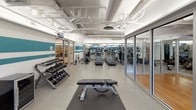 Located at 1315 W St NW in Washington, DC's vibrant Columbia Heights neighborhood, this well-appointed fitness facility offers a comprehensive workout environment across a single, thoughtfully organized floor. The space is divided into distinct functional zones, each designed to support a range of exercise modalities.

The main cardio and strength area features durable speckled flooring and is equipped with a row of treadmills, stationary bikes, and additional cardio machines. A dedicated free weights section includes a full dumbbell rack and adjustable benches, providing ample space for resistance training. The exposed ductwork and piping along the bright white ceiling lend an industrial character to the space, while teal accent stripes along the walls add a bold, energetic visual touch.

At the heart of the facility is a glass-enclosed multipurpose studio featuring warm hardwood flooring, wall-mounted ballet barres, stability balls, and a large flat-screen display — ideal for group fitness classes, yoga, or personal training sessions. The glass partitions allow natural visibility between zones while maintaining a sense of separation. Windows along the perimeter bring in natural light, enhancing the overall openness of the space. A small lounge or recovery area is also accessible, rounding out the facility's offerings.