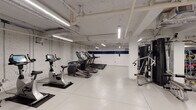 This fitness center offers a comprehensive workout environment spread across a single level. The space features an industrial aesthetic, with exposed overhead pipes, ductwork, and linear LED strip lighting running across the ceiling, complementing the white-painted brick and concrete walls accented by a bold navy blue horizontal stripe. The light-colored epoxy flooring is both durable and easy to maintain, contributing to the clean, utilitarian feel of the space.

The cardio zone is well-stocked with a row of treadmills, elliptical trainers, and stationary bikes, including True Fitness branded equipment. Adjacent to the cardio area, a dedicated strength training section houses a Cybex cable machine, a Jones functional rack, and additional cable/pulley systems. A free weights area provides a full rack of dumbbells in varying sizes alongside colorful lighter weights, medicine balls, and workout benches suited for a range of exercises.

A separate open area features a mirrored wall, exercise mats, and wall-mounted accessories, providing space for stretching, functional training, or group workouts. Sanitation stations and waste bins are conveniently placed throughout. The layout is thoughtfully organized to accommodate multiple users simultaneously while maintaining clear, open pathways throughout.