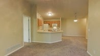This single-floor residence is designed with an open-concept layout that seamlessly connects the kitchen and living areas. The kitchen features a wraparound peninsula with a built-in double sink and light countertops, providing functional workspace and a natural division between the cooking and living zones. Warm honey-toned wood cabinetry lines the upper walls, offering ample storage, while white appliances — including a refrigerator, microwave, and range — are neatly integrated beneath. A pendant light fixture adds a subtle decorative touch above the peninsula.

The adjacent living area is carpeted in a neutral tone and anchored by a white-mantled fireplace, creating a welcoming focal point. Vaulted ceilings with a ceiling fan enhance the sense of openness and airflow throughout the space. Soft, neutral wall tones contribute to a calm and cohesive atmosphere. A sliding glass door at the far end of the living area allows natural light to filter in and provides access to the exterior. Tile flooring transitions from the kitchen area, while plush carpet covers the living space, balancing comfort with practicality throughout this well-configured home.