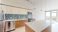 Located at 180 High Park Lane in Silver Spring, MD, this modern one-bedroom, two-bathroom residence offers a thoughtfully designed open-concept layout bathed in natural light. Light-toned hardwood flooring flows throughout, complementing the clean, contemporary aesthetic.

The kitchen is a standout feature, showcasing warm wood cabinetry paired with crisp white upper cabinets and a striking blue-green mosaic tile backsplash. A generous quartz-topped island with pendant lighting serves as both a prep space and a visual centerpiece. Stainless steel appliances, including a gas range, built-in microwave, refrigerator, and dishwasher, complete the well-appointed kitchen.

The living area opens to a private balcony through floor-to-ceiling glass doors, framing sweeping panoramic views of the surrounding treeline and skyline — filling the space with abundant sunlight throughout the day. The bedroom also enjoys direct access to the outdoors via large sliding glass doors, offering similarly expansive views and generous natural light.

Two bathrooms provide comfortable functionality; one features a soaking tub and shower combo with elegant white subway tile and a decorative mosaic accent border, finished with wood-look tile flooring. The overall design balances modern finishes with a bright, airy ambiance throughout.