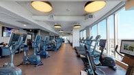 This expansive fitness center occupies a single floor at a notable elevation, offering sweeping panoramic city views through generous floor-to-ceiling windows that flood the space with natural light. The facility is thoughtfully organized into distinct zones to accommodate a wide range of workout routines.

The cardio area runs the length of the space and is lined with a comprehensive selection of equipment, including treadmills, ellipticals, and stationary bikes, all positioned to take full advantage of the striking urban vistas. Overhead, large circular pendant lights provide warm, even illumination that complements the natural light throughout the day.

A dedicated strength and functional training zone features free weights, adjustable benches, cable machines, and TRX suspension trainers, with full-length mirrors expanding the sense of space and supporting proper form. Durable flooring transitions between rubber and hard surfaces to suit each area's specific purpose.

A welcoming lounge area near the entrance is furnished with plush blue seating and a wall-mounted flat-screen TV, offering a comfortable space to relax before or after a workout. Textured wall panels and dark architectural columns add a refined, contemporary character throughout the facility.