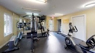This fitness center offers a comprehensive workout environment designed to accommodate a range of exercise needs. The space is laid out on a single level, with durable rubber flooring throughout that provides both safety and comfort during exercise sessions. Warm yellow-toned walls and bright fluorescent lighting create a clean, energizing atmosphere.

The room is equipped with a diverse selection of fitness equipment, including a multi-station cable weight machine, a treadmill, an upright stationary bike, a rowing machine, and a squat/barbell rack. Exercise balls and free weights are also available, making the space suitable for varied workout routines. Fitness reference charts and posters are mounted on the walls, offering guidance for users.

A large wall mirror runs along one side of the room, visually expanding the space while allowing users to monitor their form during exercise. Natural light filters in through exterior-facing windows that offer views of surrounding greenery, adding a pleasant ambiance to the workout environment. A glass-panel door provides direct exterior access. A bulletin board is mounted on the wall near an interior door, serving as a convenient communication point for residents or members. Safety features include a visible fire extinguisher and emergency exit signage. Overall, this fitness center is a practical and functional space suited for everyday fitness use.