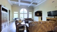 This single-floor space showcases a refined and welcoming design, highlighted by soaring coffered ceilings that lend a sense of grandeur and architectural sophistication to the main living area. The ceilings feature a crisp white grid pattern with recessed lighting, adding both style and functional illumination throughout the space.

The living area is anchored by rich, dark hardwood flooring that contrasts beautifully with the warm, neutral-toned walls. A set of generously sized leather sofas and traditional wooden accent tables create a comfortable and classic seating arrangement. The focal point of the room is a stunning arched window flanked by tall rectangular windows, flooding the interior with an abundance of natural light and providing views of the surrounding greenery.

An elegant arched entryway leads to an adjacent room finished in a deep, contrasting wall color, adding a dramatic yet cohesive design element. This secondary space appears to function as a study or office area, complete with built-in cabinetry and warm wood finishes.

Throughout the space, careful attention to architectural detail is evident — from the decorative crown molding to the classic column accents — creating an environment that is both timeless and inviting.