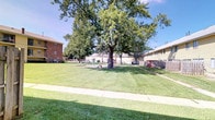 This single-floor property is distinguished by its expansive shared outdoor grounds, which serve as a central community feature. The exterior landscape is anchored by several large, mature trees that provide generous shade and a well-established, park-like atmosphere. The grounds are covered in neatly maintained green lawn that extends across a wide open area, offering ample space for outdoor activities and relaxation.

A dedicated playground area is visible within the grounds, equipped with play structures, making the space particularly well-suited for families with children. Concrete walkways traverse the lawn, providing clean and accessible pathways throughout the outdoor area.

The surrounding buildings feature a mix of classic brick construction and light-colored siding exteriors, contributing to a traditional residential character. Fencing sections define portions of the property boundary, adding a sense of privacy and organization to the outdoor spaces.

The overall ambiance of the property is open, green, and community-oriented, with the mature tree canopy and well-kept grounds creating a welcoming outdoor environment. Under clear blue skies, the natural landscaping and communal amenities combine to offer a comfortable and functional setting for residents.