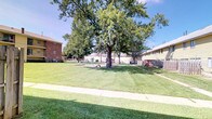This single-floor property is distinguished by its expansive shared outdoor grounds, which serve as a central community feature. The exterior landscape is anchored by several large, mature trees that provide generous shade and a well-established, park-like atmosphere. The grounds are covered in neatly maintained green lawn that extends across a wide open area, offering ample space for outdoor activities and relaxation.

A dedicated playground area is visible within the grounds, equipped with play structures, making the space particularly well-suited for families with children. Concrete walkways traverse the lawn, providing clean and accessible pathways throughout the outdoor area.

The surrounding buildings feature a mix of classic brick construction and light-colored siding exteriors, contributing to a traditional residential character. Fencing sections define portions of the property boundary, adding a sense of privacy and organization to the outdoor spaces.

The overall ambiance of the property is open, green, and community-oriented, with the mature tree canopy and well-kept grounds creating a welcoming outdoor environment. Under clear blue skies, the natural landscaping and communal amenities combine to offer a comfortable and functional setting for residents.