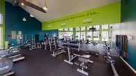 This fitness center is a well-appointed, single-floor facility designed to support a comprehensive workout experience. The space is immediately distinguished by its soaring vaulted ceiling with exposed wooden beams and visible ductwork, lending an industrial-chic aesthetic balanced by bold, energizing colors. The walls are painted in striking teal and lime green, with motivational phrases such as "Everyday is another chance to be the best version of you..." prominently displayed along the upper green wall, reinforcing the facility's encouraging atmosphere.

Floor-to-ceiling windows line multiple walls, flooding the interior with generous natural light while offering views of lush greenery and the surrounding outdoor landscape. The dark rubber flooring provides a practical, durable surface throughout the space.

The cardio zone features a row of treadmills, ellipticals, and stationary bikes positioned along the windowed walls, complemented by a wall-mounted flat-screen television. A dedicated strength training area is equipped with free weights, dumbbells, barbells, weight benches, and resistance machines. A rowing machine and spin bike round out the equipment selection. The entrance area includes a sanitization station, adding a functional and hygienic welcome point to the facility.