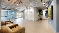 Toluca Court presents a thoughtfully designed single-story communal and leasing space that combines contemporary aesthetics with functional layout. The lobby is anchored by a striking three-dimensional white textured accent wall bearing the Toluca Court name, serving as an immediate focal point upon entry. Exposed ductwork and conduit traverse the ceiling, lending an industrial character that contrasts with the smooth, large-format light-toned tile flooring found throughout.

The entry area opens into a spacious reception and lounge zone, where velvet seating in warm amber tones adds a welcoming residential quality. Pendant lighting and adjustable track lighting provide layered illumination across the open floor plan. Large windows along the perimeter allow generous natural light to filter in, offering views of surrounding greenery.

A glass-walled leasing office with frosted tile detailing at its base provides a semi-private workspace while maintaining visual connectivity to the broader space. Inside, workstations are bathed in natural light from multiple windows. Lush indoor plants are positioned throughout, including bird of paradise and snake plants, enhancing the fresh, airy ambiance. Colorful artwork displayed on blue accent walls adds vibrancy and personality to the overall environment.