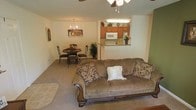 This single-floor space features an open-concept design that seamlessly integrates the living, dining, and kitchen areas into one cohesive layout. The living room is anchored by a generously sized, plush sofa with decorative pillows, complemented by soft wall-to-wall carpeting that adds warmth underfoot. A ceiling fan overhead provides both comfort and lighting for the space.

Adjacent to the living area, the dining space accommodates a round table with seating for multiple guests, lit by an elegant chandelier that adds a touch of classic style to the room. A decorative plant nearby adds a natural element to the setting.

The kitchen is accessible via a pass-through opening in the wall, allowing visual connectivity between spaces. Fitted with warm wood-toned cabinetry, a built-in microwave, a refrigerator, and a raised countertop bar that doubles as a casual dining or serving surface, the kitchen is both practical and inviting.

Throughout the space, neutral-toned walls and consistent carpeting create a unified, calm ambiance, making the overall layout feel open, approachable, and well-suited for everyday living.