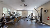 Located at 8800 Dunwoody Pl in Sandy Springs, GA, this fitness center offers a well-organized and comprehensively equipped workout facility spread across a single floor. The space features neutral gray walls, recessed lighting, and durable rubber flooring designed to withstand heavy use while providing comfort and safety during exercise.

The main workout area is generously sized, housing a row of cardio machines that includes treadmills, ellipticals, and other aerobic equipment. A ceiling fan aids in air circulation, complementing the HVAC system to maintain a comfortable environment during intense workouts. Multiple large windows line the walls, flooding the space with natural light and offering views of the surrounding greenery.

Strength training is well-represented, with a multi-station cable machine, a dedicated weight bench, and a rack stocked with an assortment of dumbbells and kettlebells in varying weights. A stability ball and exercise mat provide options for functional and flexibility training. An additional room or alcove extends the usable space, housing further strength equipment including a leg extension machine and a power rack with an adjustable bench.

A glass-paneled exit door provides convenient outdoor access. The facility also includes a fire extinguisher and other safety provisions, reflecting a thoughtfully maintained environment. Overall, this fitness center delivers a clean, bright, and functional space suited to a wide range of training needs.